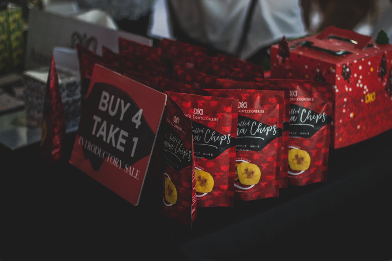 Packaged banana chips displayed with a promotional Buy 4 Take 1 sign at an indoor sale event.